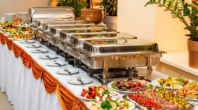 A long buffet table with chafing dishes, plates, and various appetizers and salads.