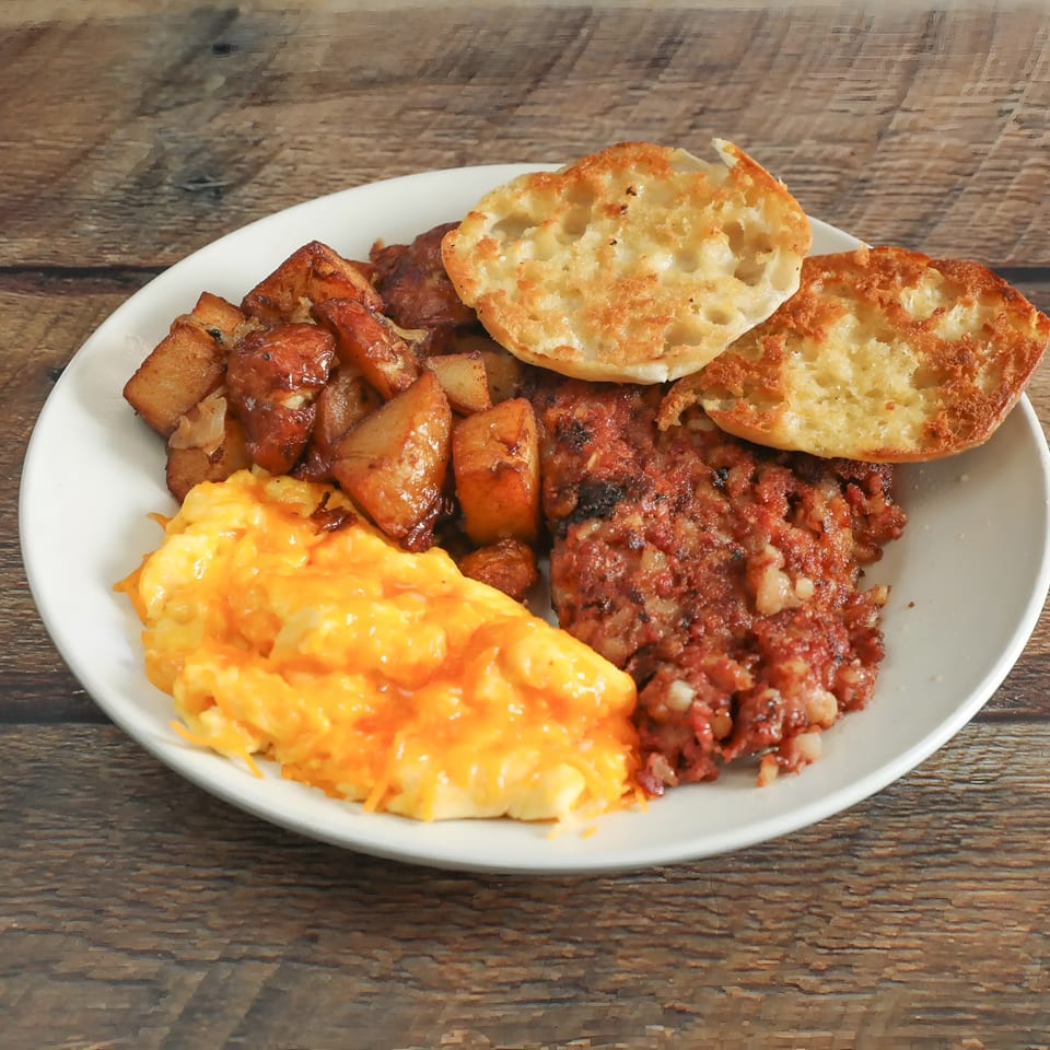 A breakfast plate with cheesy scrambled eggs, home fries, corned beef hash, and an English muffin.