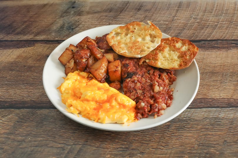 A breakfast plate with cheesy scrambled eggs, home fries, corned beef hash, and an English muffin.