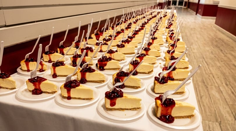 Numerous cheesecake slices with cherry topping and plastic forks arranged on a long table.