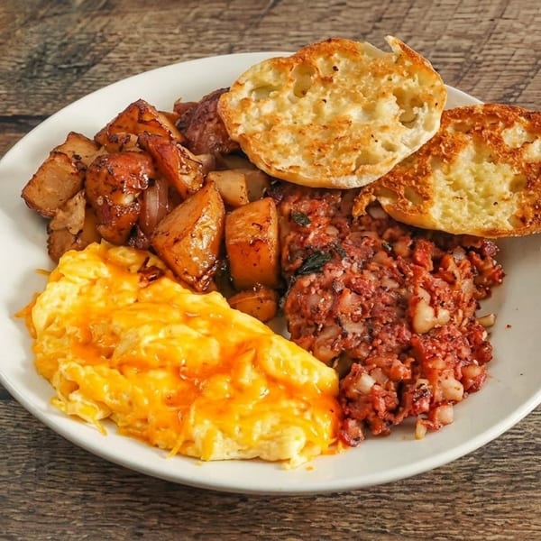 A breakfast plate with cheesy scrambled eggs, home fries, corned beef hash, and an English muffin.