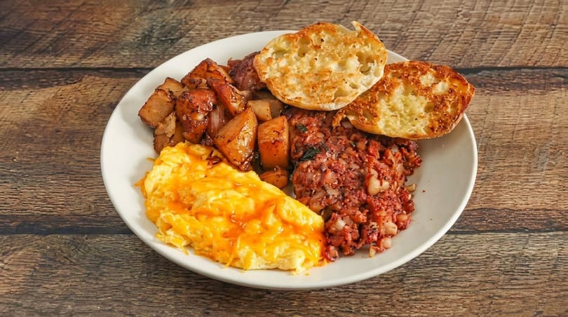 A breakfast plate with cheesy scrambled eggs, home fries, corned beef hash, and an English muffin.