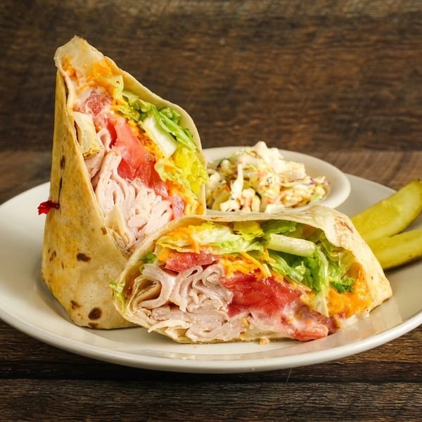 Two halves of a turkey wrap with lettuce, tomato, and cheese, served with coleslaw and pickles.
