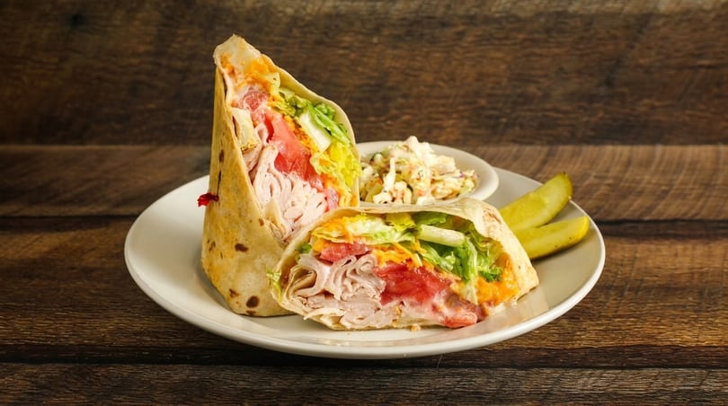 Two halves of a turkey wrap with lettuce, tomato, and cheese, served with coleslaw and pickles.