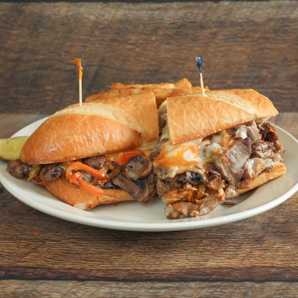 Two steak and cheese subs with mushrooms, peppers, and onions, served with fries and a pickle.