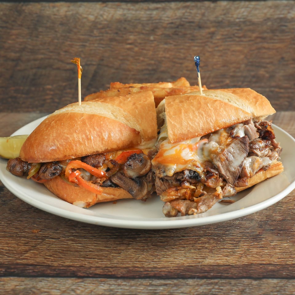 Two steak and cheese subs with mushrooms, peppers, and onions, served with fries and a pickle.
