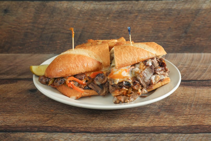 Two steak and cheese subs with mushrooms, peppers, and onions, served with fries and a pickle.