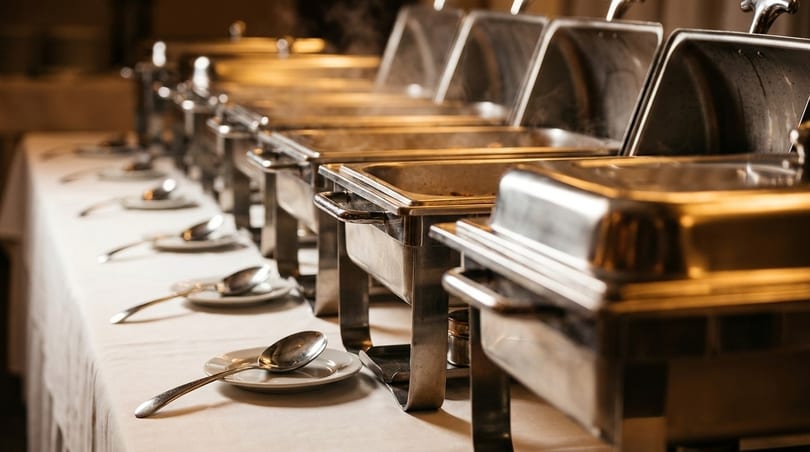 A buffet line with several steaming chafing dishes and serving spoons on a white tablecloth.