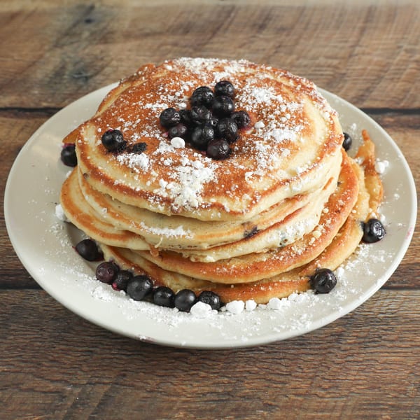 Stack of blueberry pancakes dusted with powdered sugar on a white plate, on a wooden table.