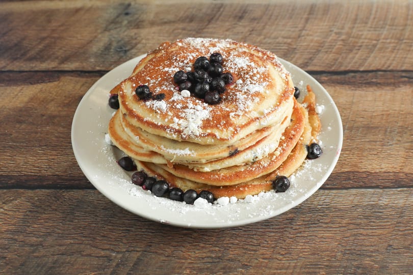 Stack of blueberry pancakes dusted with powdered sugar on a white plate, on a wooden table.