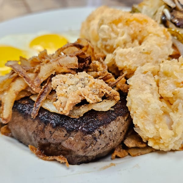 Steak with fried onions, shrimp, and two sunny-side-up eggs on a white plate.