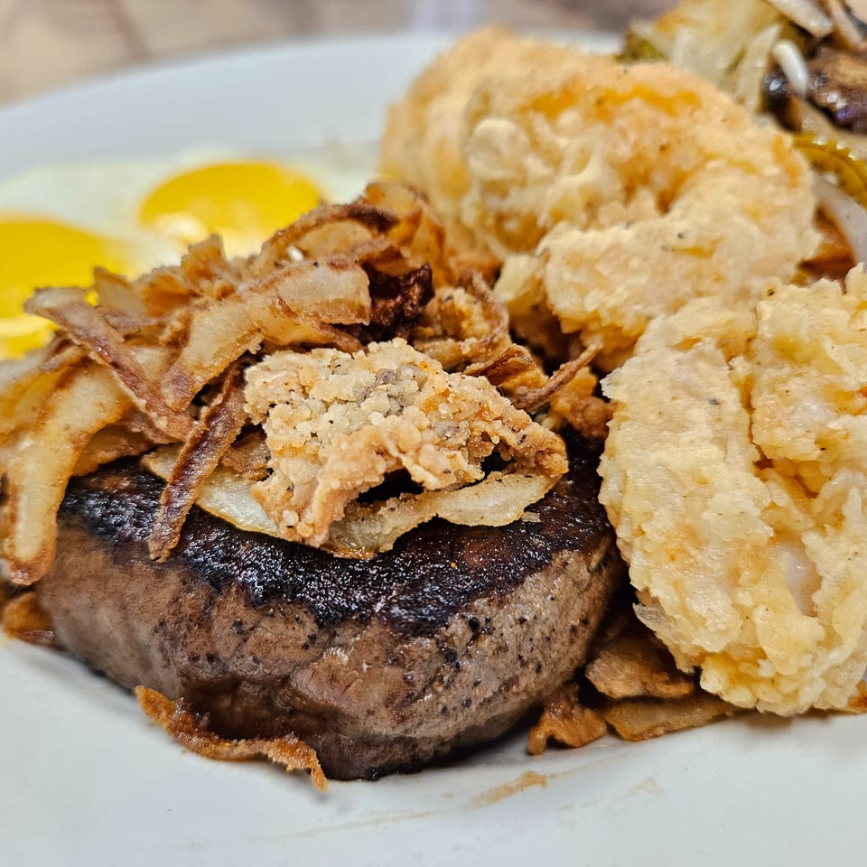 Steak with fried onions, shrimp, and two sunny-side-up eggs on a white plate.