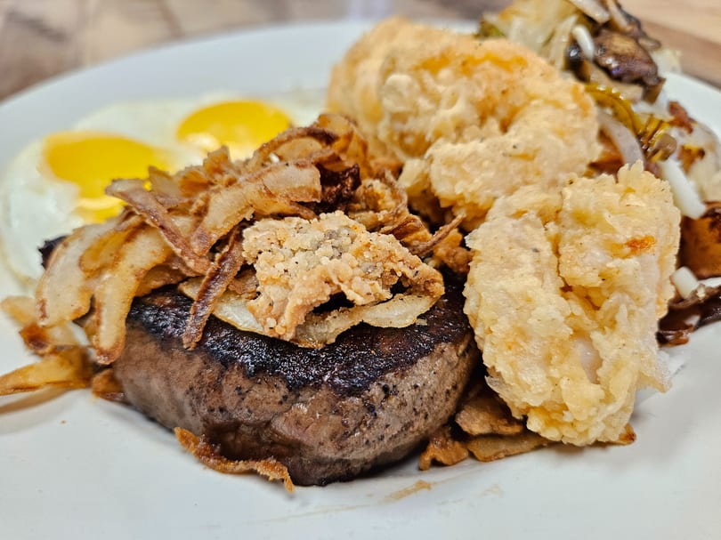 Steak with fried onions, shrimp, and two sunny-side-up eggs on a white plate.