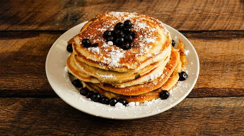 Stack of golden pancakes topped with blueberries and powdered sugar on a white plate.